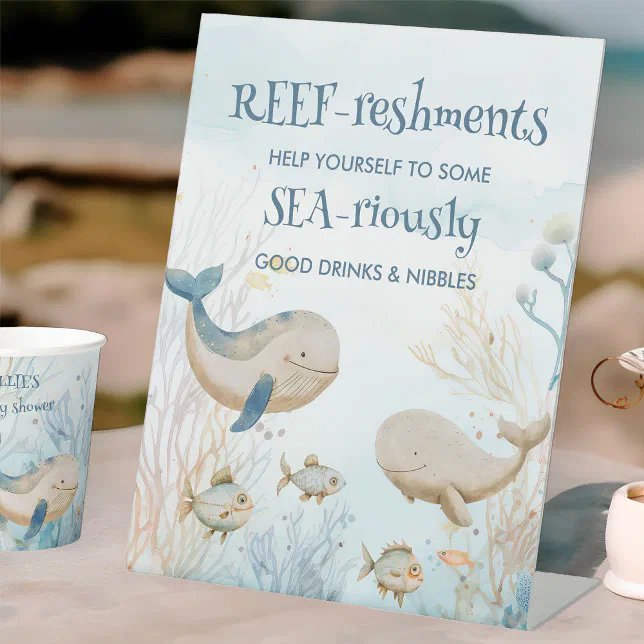 A playful sea-themed baby shower pedestal sign is placed on a table to direct guests to the drink and food stations, with a paper cup in the same design placed nearby.