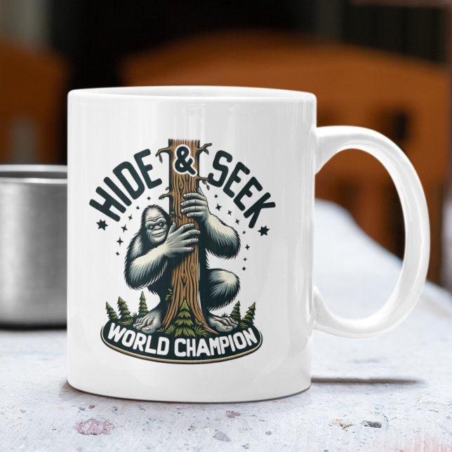 Funny Hide & Seek World Champion Bigfoot Coffee Mug (White mug with Bigfoot "Hide & Seek World Champion" design on an outdoor table.)