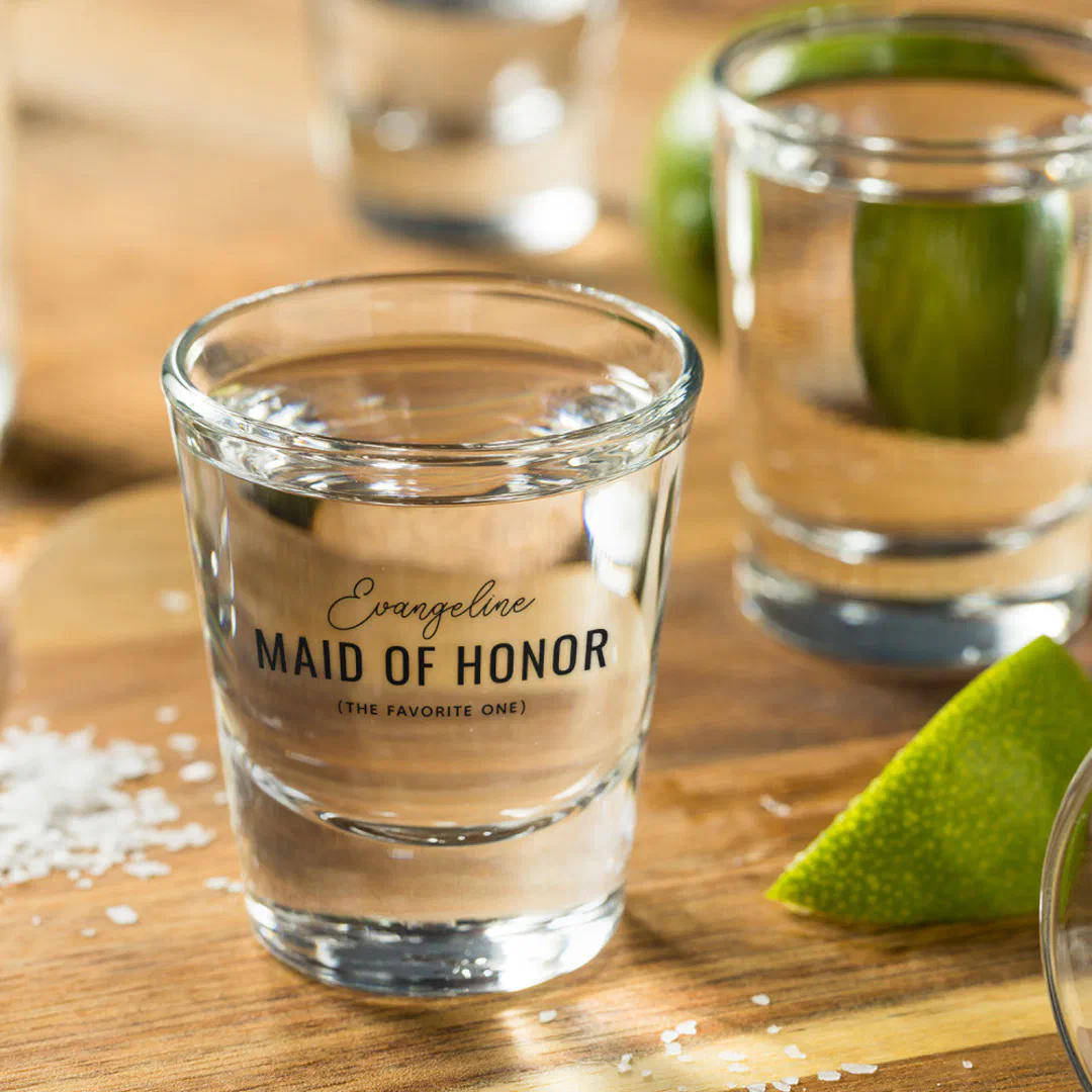 A clear shot glass with black script. There's a chopping board, lime and salt in the backrgound.