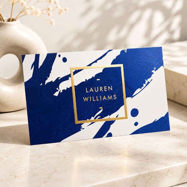 A rectangular business card featuring a bold, textured deep indigo blue brushstroke pattern against a white background, highlighted by a metallic gold foil square frame with a professional name in gold foil letters.