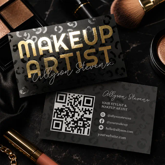 Two black foil business cards with a subtle, glossy leopard print texture are artfully arranged on a dark marble surface alongside cosmetics, including a lip gloss tube and a large powder brush.