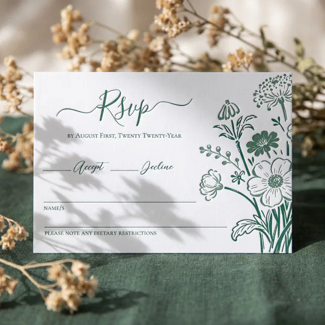 A green letterpressed RSVP card lying on a textured green material with foliage in the background and foreground.