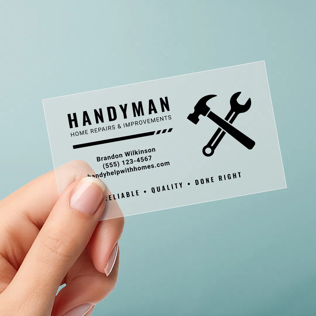 A hand with manicured nails is holding up a clear business card with black text. The background is neutral.