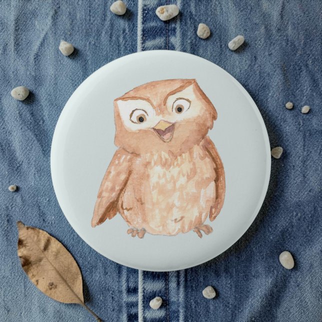 Happy Owl Watercolor Button (A watercolor owl cartoon smiling on a pale blue pin, laid on denim amid pebbles and leaves.)