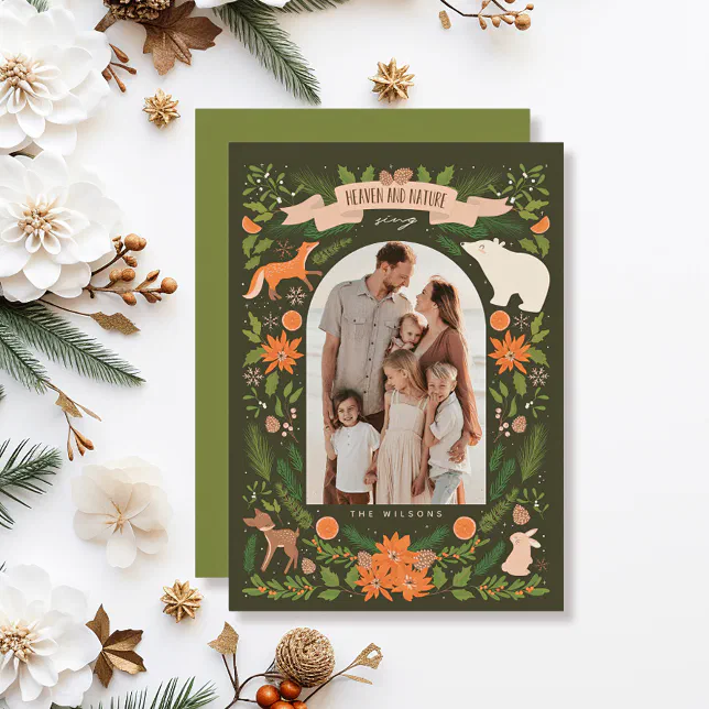 A green holiday card illustrated with woodland creatures surrounded by white flowers, pine cones, and fir tree sprigs.
