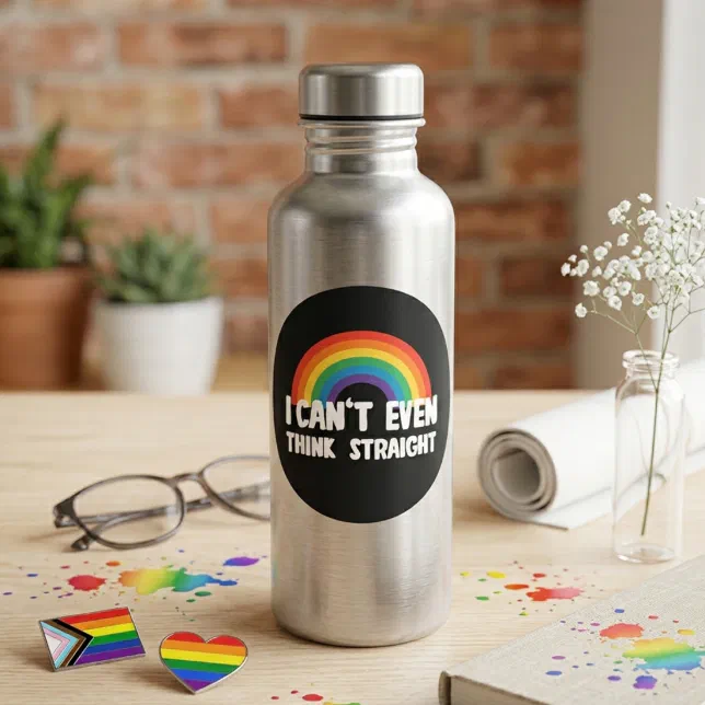 A Pride-themed sticker is placed on a metallic water bottle, with plants and a brick wall in the background. Glasses, Pride pins and a flower are pictured scattered on the table around the bottle. 