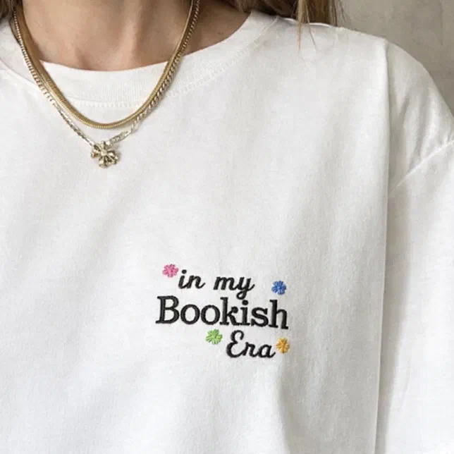A person is wearing an embroidered book club-inspired sweatshirt and gold necklaces.