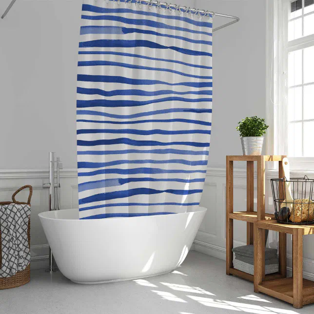 A blue and white striped shower curtain shown in use in a bathroom. In the background there is plants on a brown shelving unit and a wash basket.