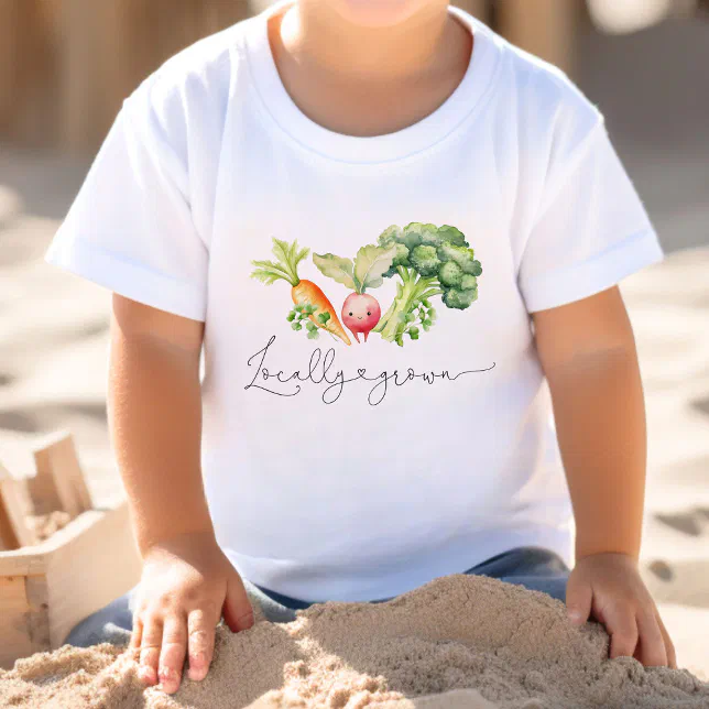 A child sitting in sand wearing a white tshirt with a carrot, radish and a large stemmed broccoli.