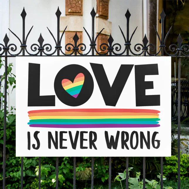 A Pride-themed sign is placed on a black fence, with bushes and the entrance to a house in the background.