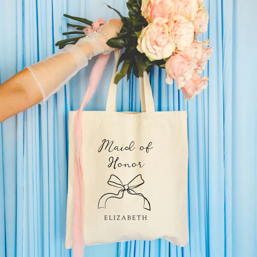 Gloved hand holding flowers and Maid of Honor coquette bow tote bag in customizable colors on a blue background.