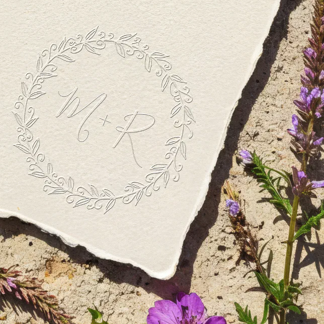 A wedding embosser print with a floral border and the couple's initials. Wildflowers are scattered near the embossed material.