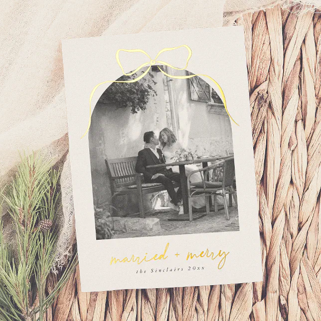 A white and gold holiday card resting on a woven surface next to some greenery.
