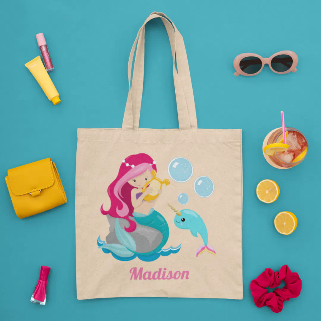 A natural canvas tote bag laid flat on a bright teal surface, printed with an illustrated pink-haired mermaid seated on a rock with a smiling narwhal and floating bubbles, surrounded by a yellow mini wallet, a tube of lip gloss, pink nail polish, a pink scrunchie, oval peach sunglasses, a halved lemon, and an iced drink with a pink straw and lemon garnish.