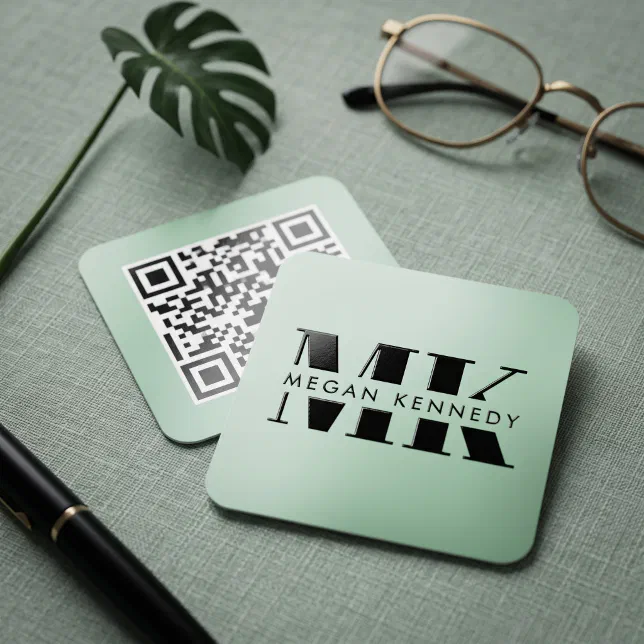 A square mint green business card is lying on a green surface, next to a pen, a leaf, and a pair of glasses.