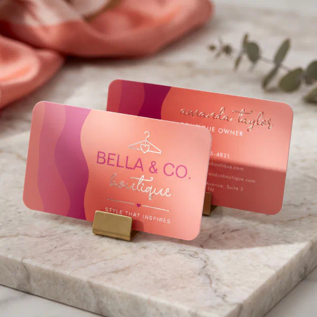 A modern, rounded-corner business card stands upright in brass clips on a white marble surface, displaying a vibrant pink, magenta, and peach gradient background with elegant rose gold foil lettering.
