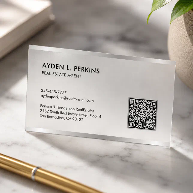 A frosted business card with a QR code sits on a marble surface with a pen in front of it and a plant behind it.