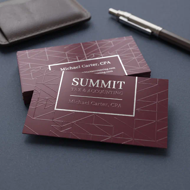 Premium, matte burgundy business cards sit on a dark gray surface next to a brown leather wallet and a sleek pen. The card features a Spot UV geometric line pattern with a central, rectangular silver foil border that frames the text.