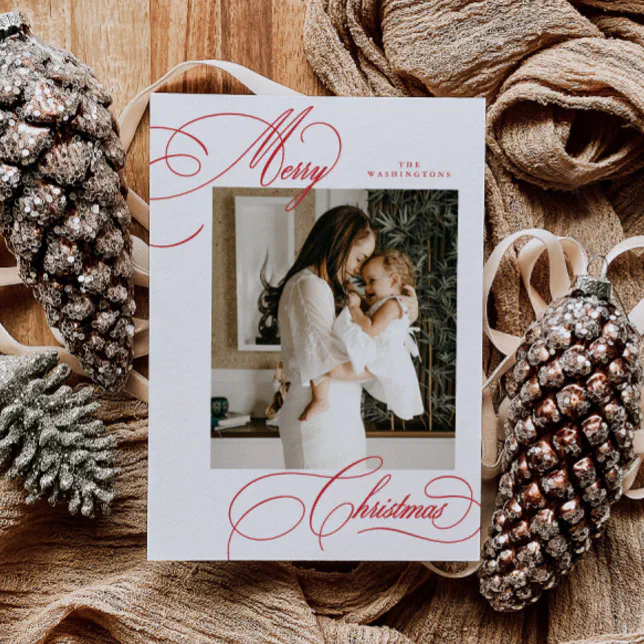 A red script holiday card surrounded by pine cones and rustic brown fabric.