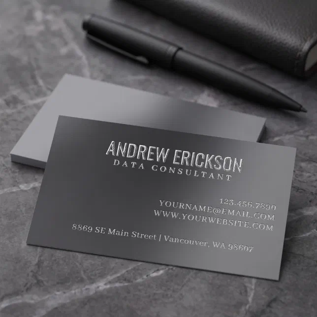 A charcoal-colored business card is lying next to a black pen on a marble surface.
