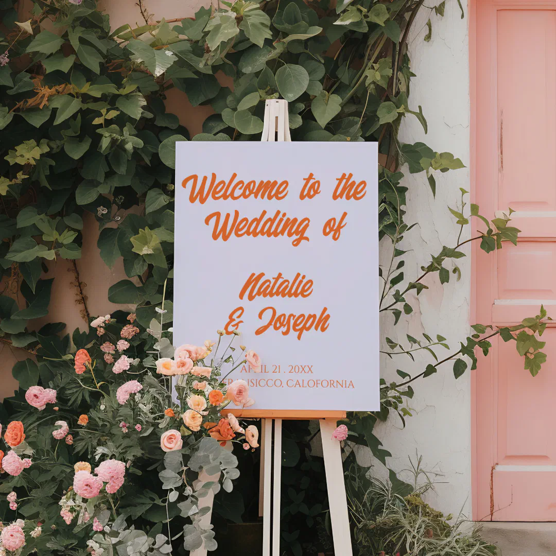 A wedding welcome sign with bold retro orange script, displayed on a wooden easel surrounded by climbing greenery and pink and peach flowers beside a pastel pink door.
