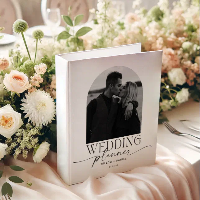 A wedding planner binder is placed on top of a soft pink velvet cloth, with blush colored flowers and greenery elegantly placed in the background.