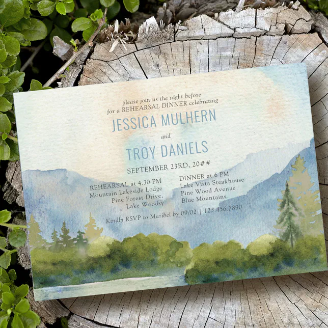 A rehearsal dinner invitation with watercolor artwork depicting a mountain range and lake. The invite is lying on a tree stump.