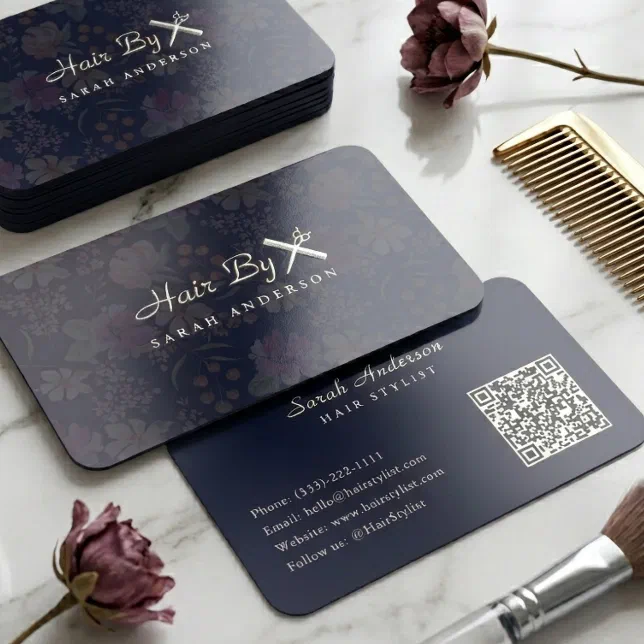 A dark navy blue business card with silver foil design lies face up on a light marble surface, surrounded by styling accessories.