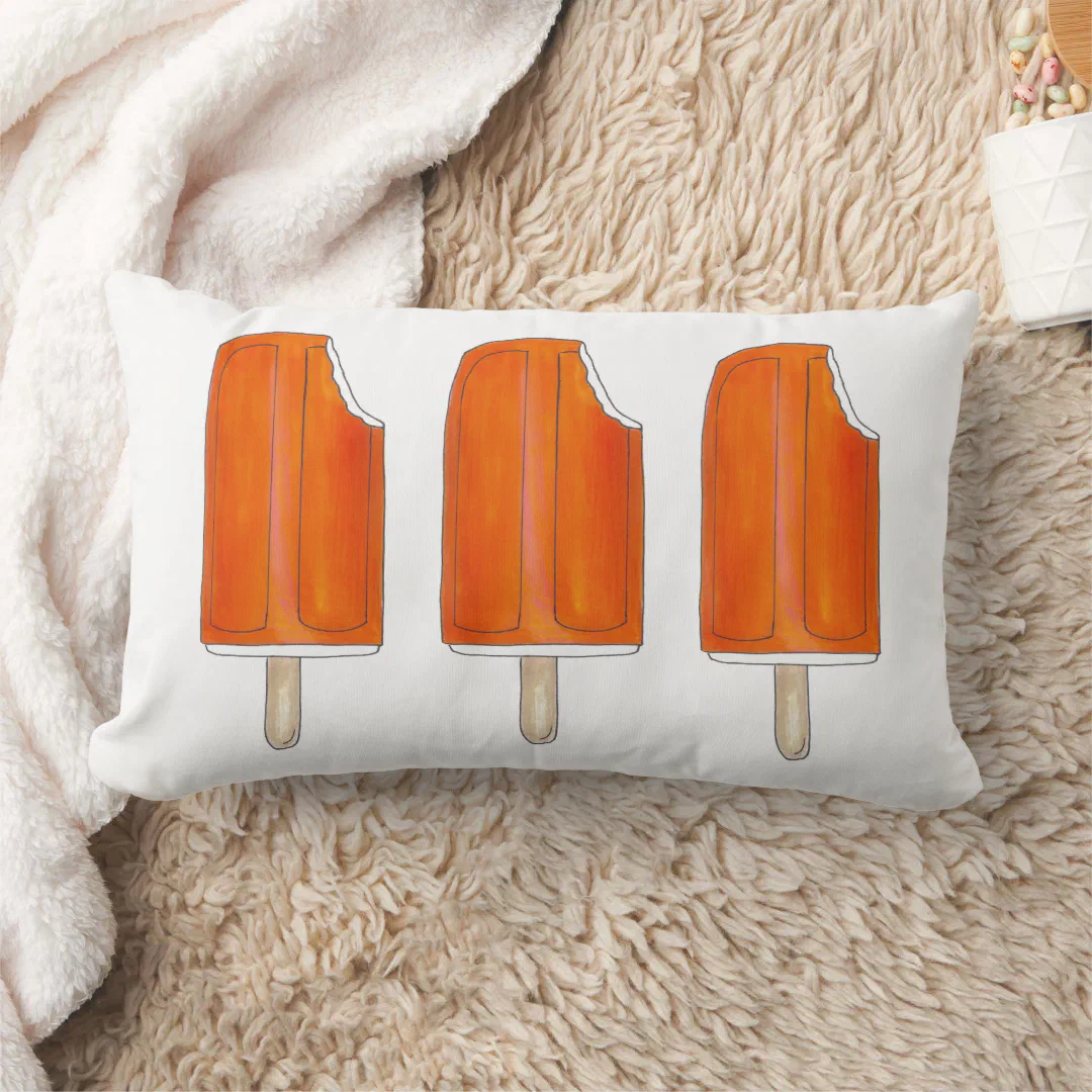 In the back of the image there's a cream fleece blanket to the left and a cream matted rug in the main frame. In the center is a white pillow with 3 mildly-bitten orange popsicles printed.