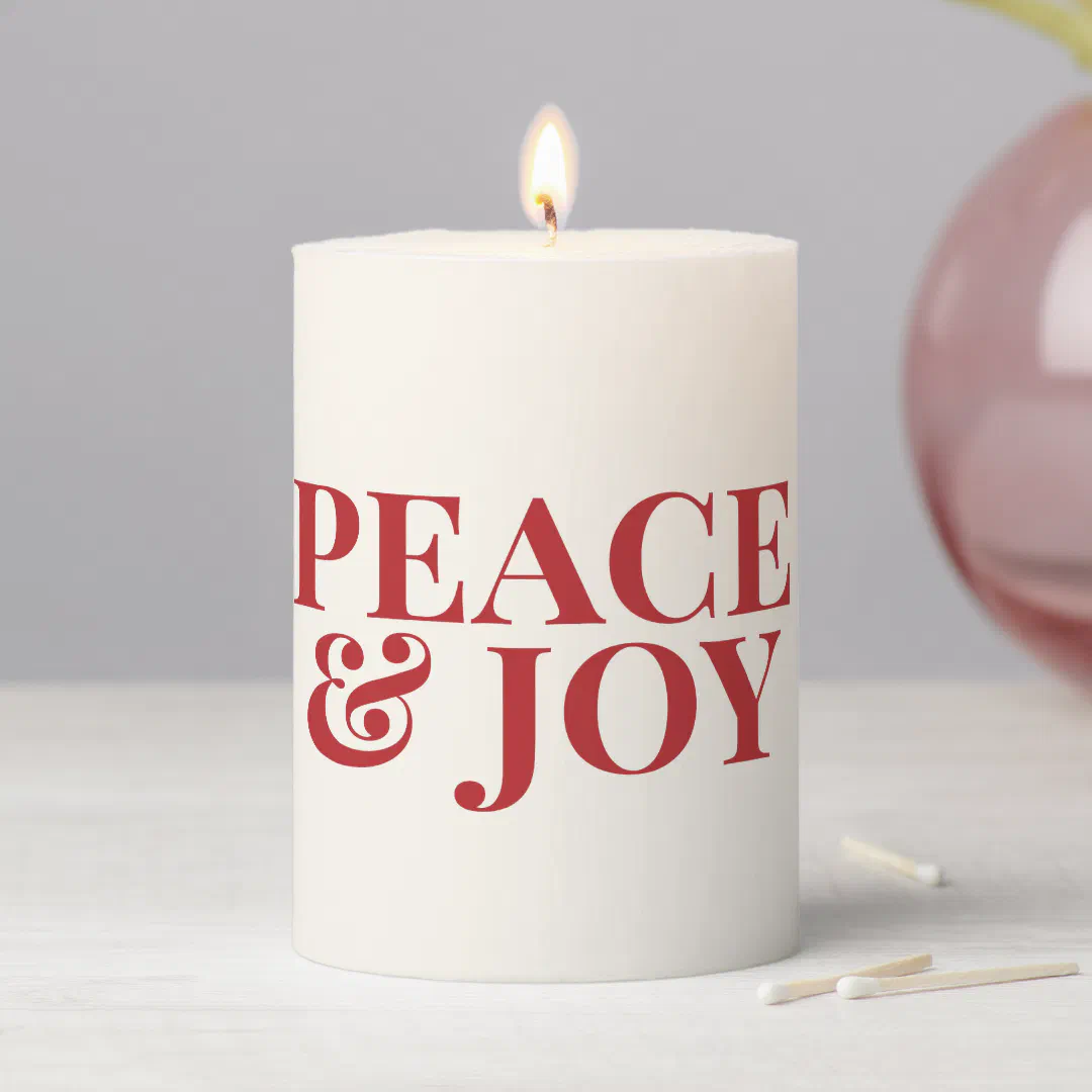 A lit cream pillar candle printed with bold red serif lettering, sitting on a whitewashed wood surface with two matchsticks and a pink glass vase against a gray wall.