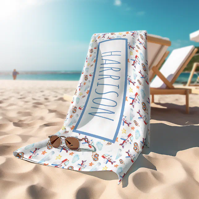 The background is of a beach with white loungers, The white towel has cartoon aeroplanes printed on them and the personalized name in a big classic font in the shade of blue. Theres a pair of sunglasses at the edge of the towel.