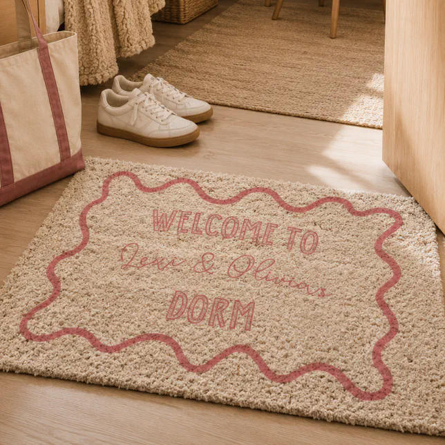A dorm room welcome mat that has "Welcome to Lexi & Olivia's dorm" printed on it. There's a tote bag and a pair of shoes in the background.