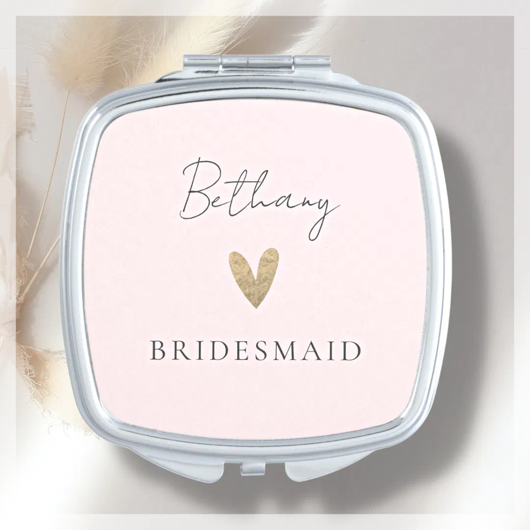 A pale white compact mirror with a silver rim. On the front there's a scripted name and a faux-foil gold heart. In the background theres various shades of cream and gray.