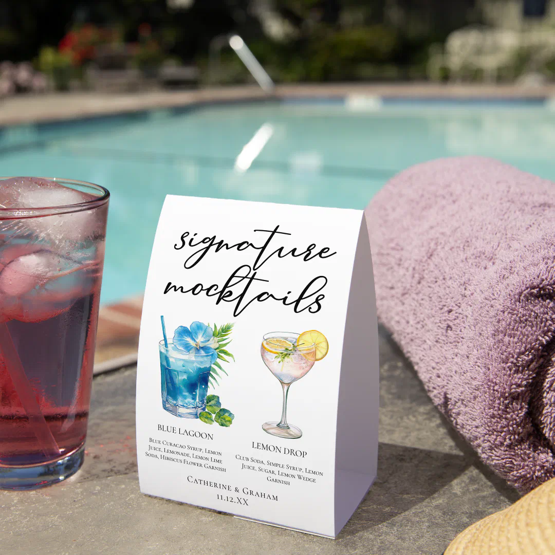 A folded white menu card propped on a concrete pool edge beside a chilled iced drink in a clear highball glass, a loosely rolled purple towel, and the edge of a natural straw hat, with a sunlit aqua pool and soft garden backdrop out of focus behind it.