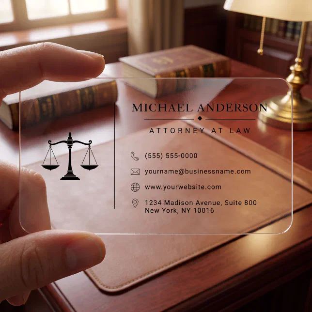 A person holds a clear business card in front of a lawyer's desk. Books and a lamp can be seen behind the card.
