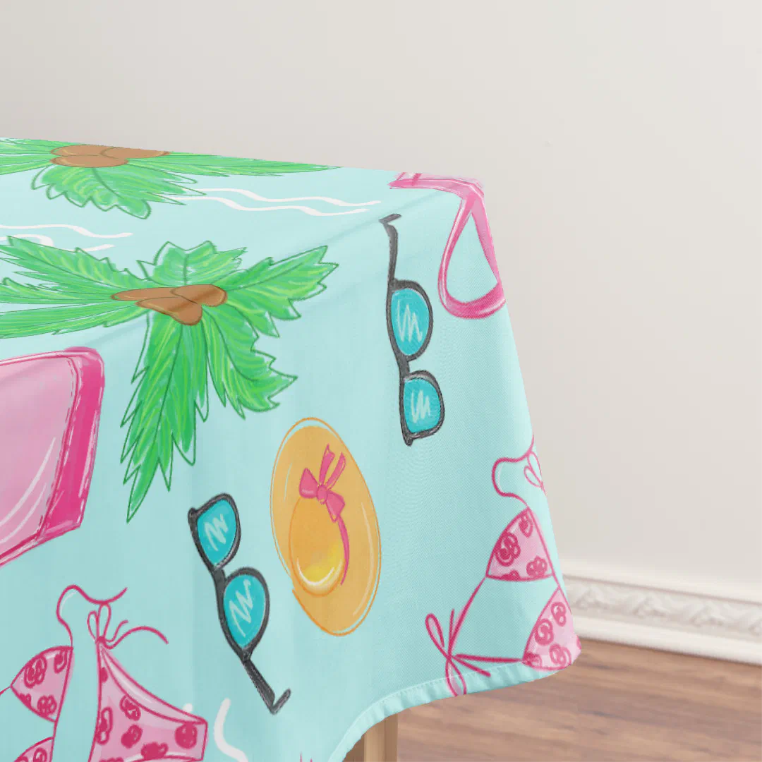 A mint green tablecloth on a table against a plain white wall, printed with a playful hand-drawn pattern of tropical and beach accessories including oversized pineapples, pink shopping bags, retro sunglasses, a yellow sunhat with a bow, and pink spotted swimsuits scattered across the surface.