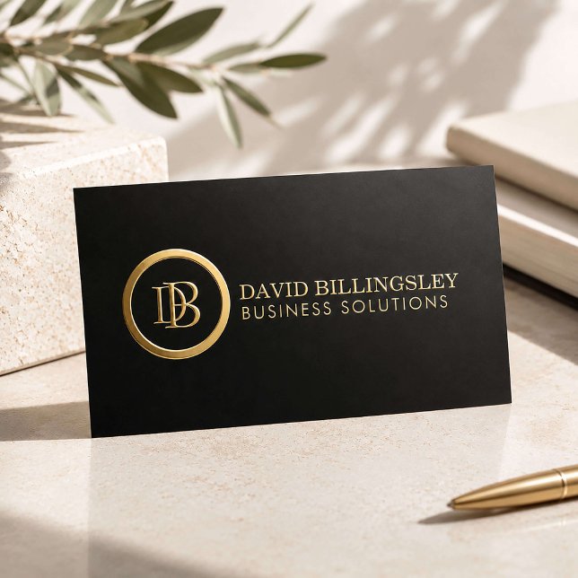 A minimalist black matte business card features a striking metallic gold foil circular monogram emblem on the left with professional details in clean, elegant gold foil letters.