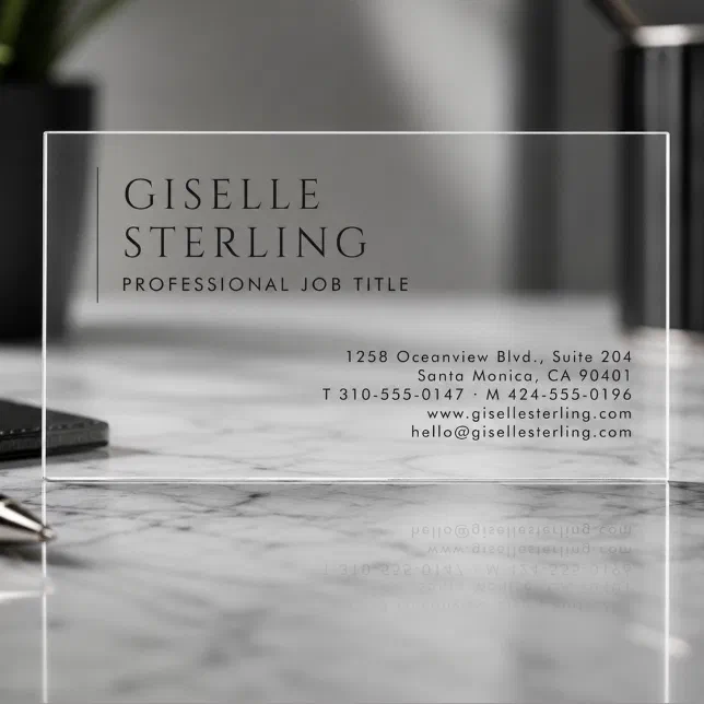 A clear business card is standing on a grey marble desk, with a pen lying against it.