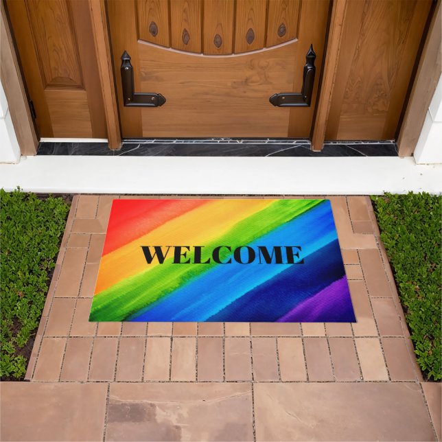 Rainbow welcome doormat set down in front of a brown door. In the background there is a grass lawn.