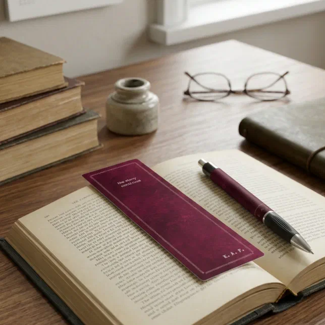 A burgundy bookmark is shown being used inside a book on a table next to a pen, a pair of glasses and some other books.