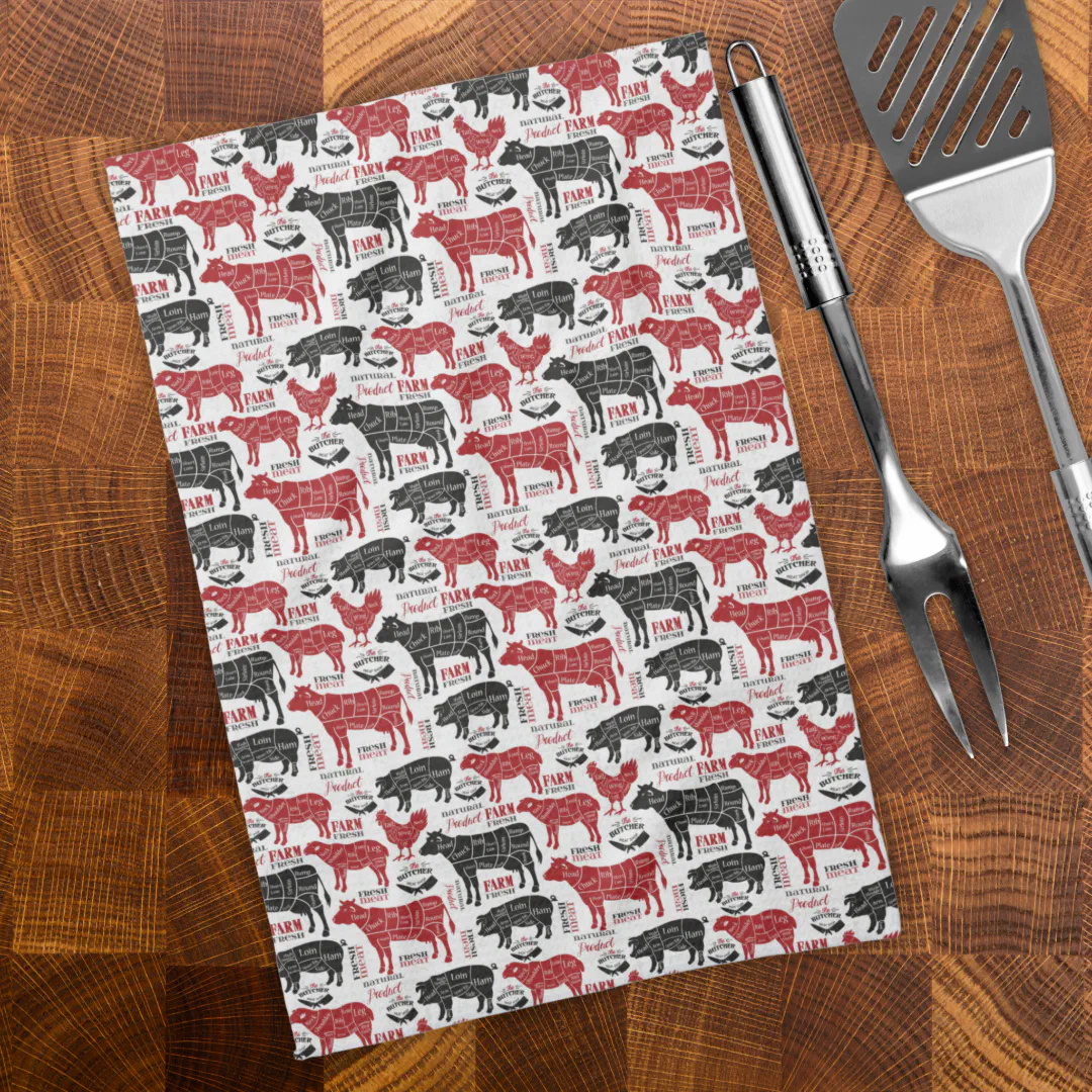 A dark-wooden background with a white kitchen towel as the main display. There are black and red clip art images of farm animals on the towel. To the right there's silver bbq utensils.