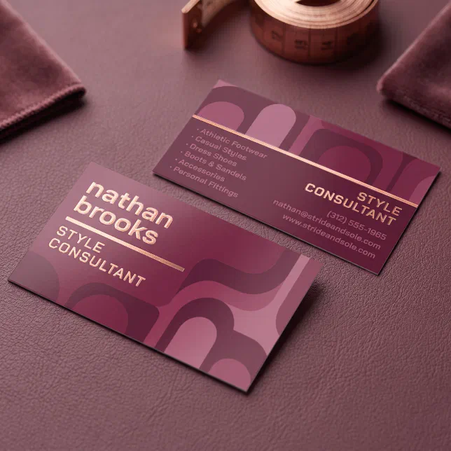 A modern, retro-inspired geometric patterned business card with rose gold foil and wavy shapes in shades of deep burgundy, mauve, and dusty rose. It sits on a dark wine red surface, with a metallic copper measuring tape and matching fabric samples.