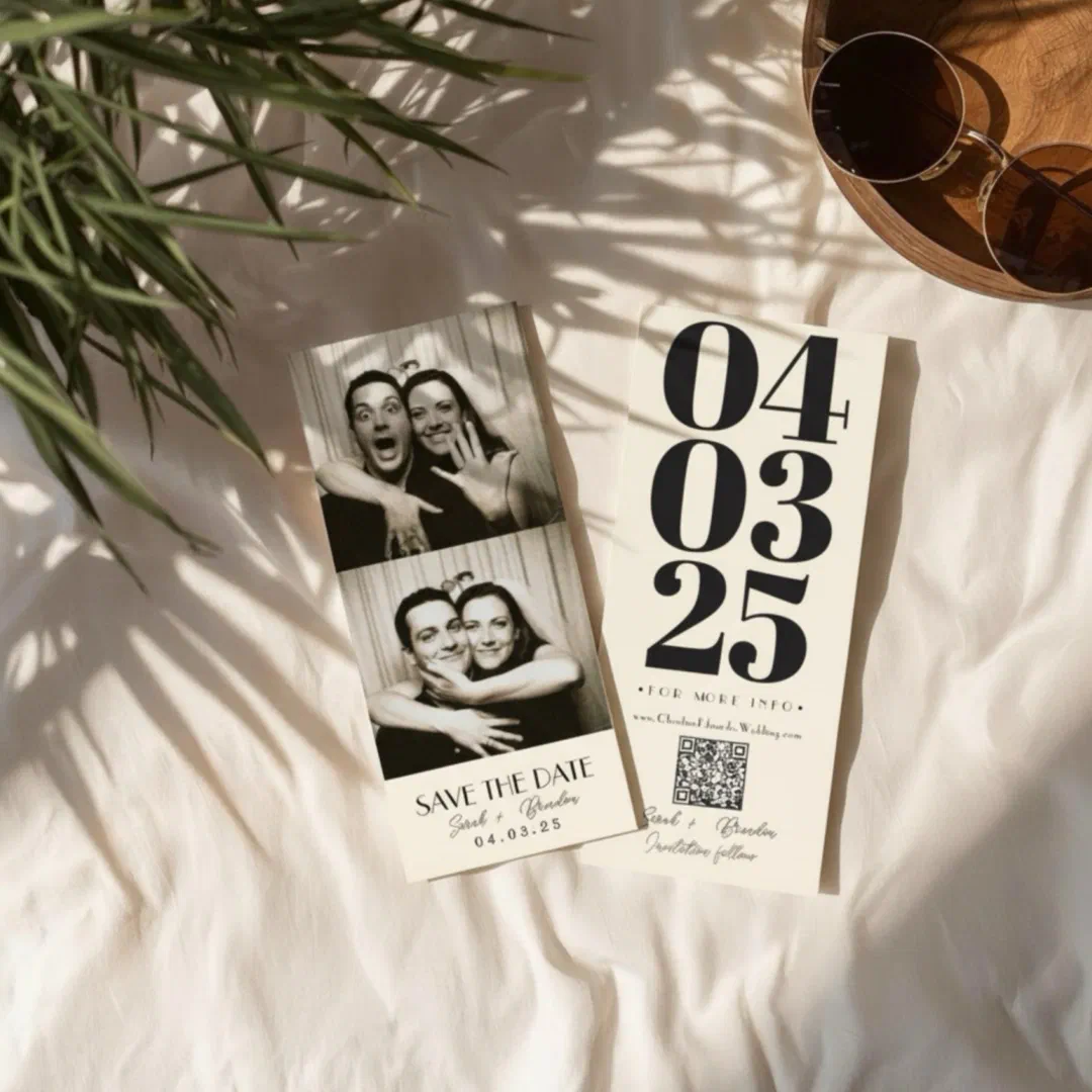 A retro photo booth style save the date featuring black and white couple photo strips, shown front and back with bold vintage date typography, laid on white bedding beside sunglasses on a wooden tray and leafy shadows.