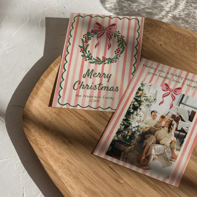 A pink and white striped holiday card with a wreath and bow design is seen on a wooden plate.