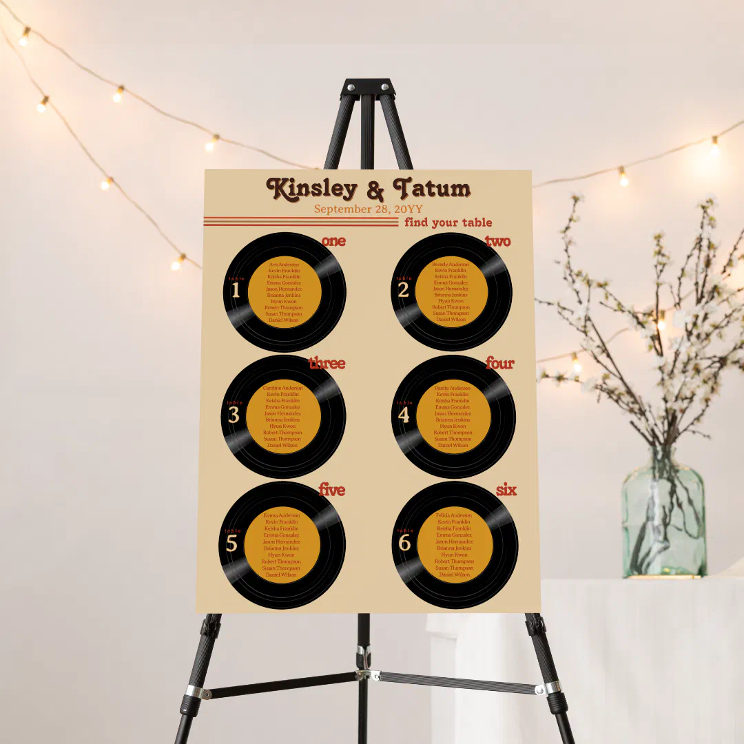 A wedding seating chart foam board displaying guest names on six illustrated vinyl records, set on a black easel with string lights and blossom branches in the background.