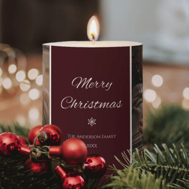 A dark red Christmas-themed candle with Christmas tree branches and red décor in the front set against a background of blurred string lights.