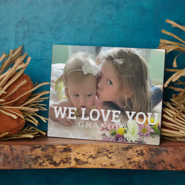A plaque for a special grandfather featuring a young girl kissing her baby sister. Some foliage can be seen in the background, and the plaque is on a wooden mantel. 