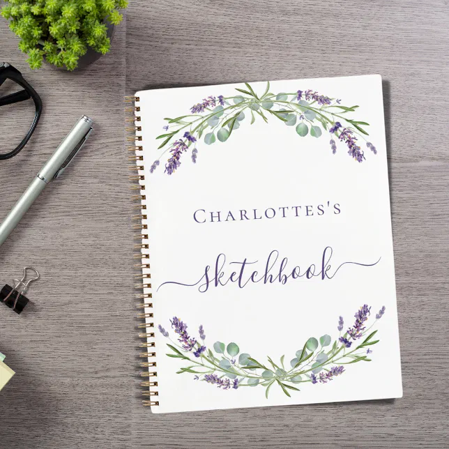 A white sketchbook with purple lavender and greenery sits on a grey desk next to a plant, a pair of glasses, a paper clip and a pen.