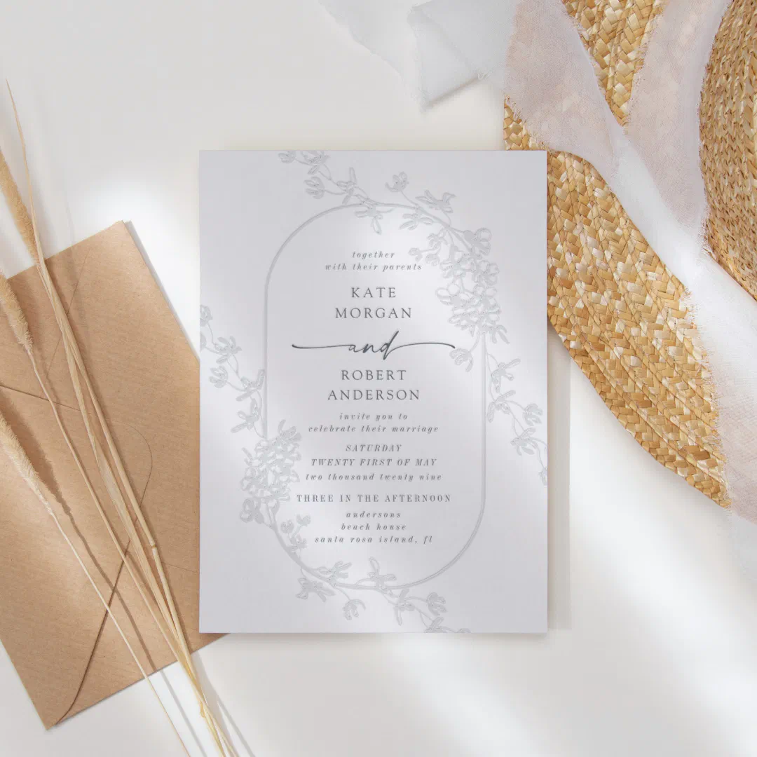 The invitation design illustrates a clear round border around the text with leaf branches hanging off the top and bottom. In the background there's a beach hat, brown envelope and twigs.