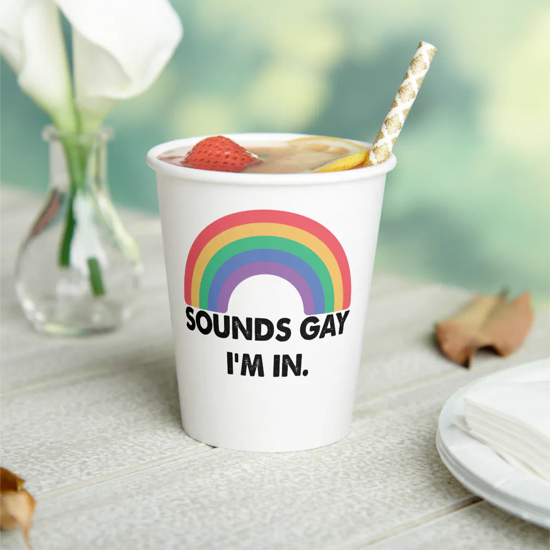White paper cup with rainbow motif. In the background there is a vase with a white flower, a plate, napkins, and some leaves.